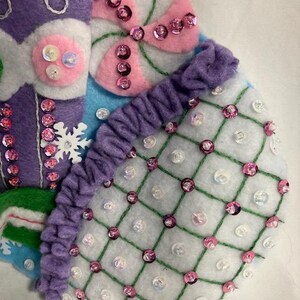 May include: Close-up of a handmade Christmas stocking with felt and sequin embellishments. The stocking features a purple section with white stitching, a pink and white candy design, and a white section with a grid pattern and pink sequins. The design is festive and colorful.
