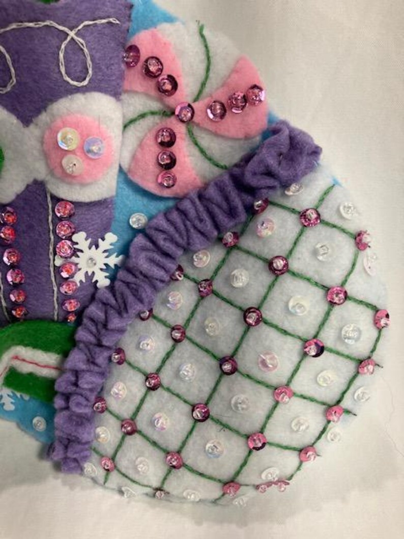 May include: Close-up of a handmade Christmas stocking with felt and sequin embellishments. The stocking features a purple section with white stitching, a pink and white candy design, and a white section with a grid pattern and pink sequins. The design is festive and colorful.