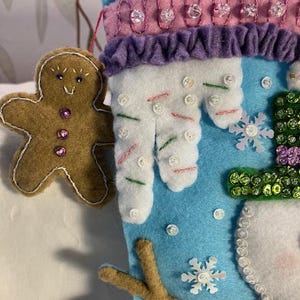 May include: A handmade Christmas stocking with a snowman design. The stocking is blue, white, and purple, with a gingerbread man ornament. The snowman has a green hat and a pink mouth. The stocking is decorated with sequins and buttons.