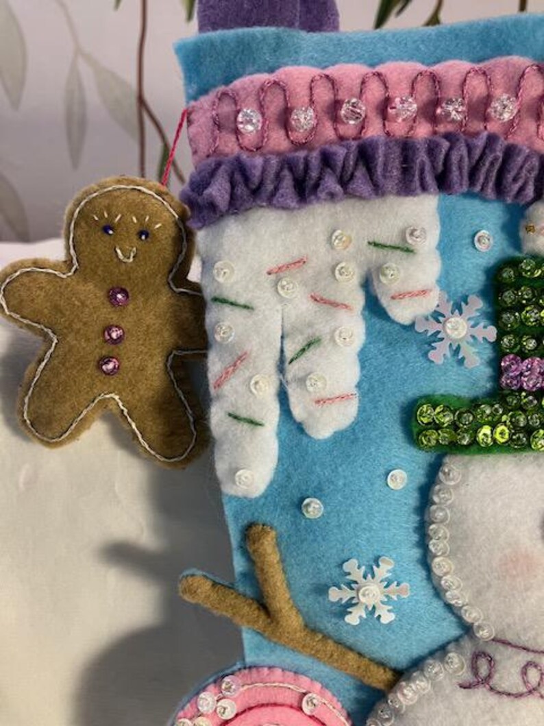 May include: A handmade Christmas stocking with a snowman design. The stocking is blue, white, and purple, with a gingerbread man ornament. The snowman has a green hat and a pink mouth. The stocking is decorated with sequins and buttons.