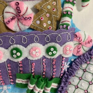 May include: Close-up of a felt Christmas stocking with candy and gingerbread decorations. The stocking features a purple cupcake design, a green and white candy cane, and a gingerbread tree, all embellished with sequins.