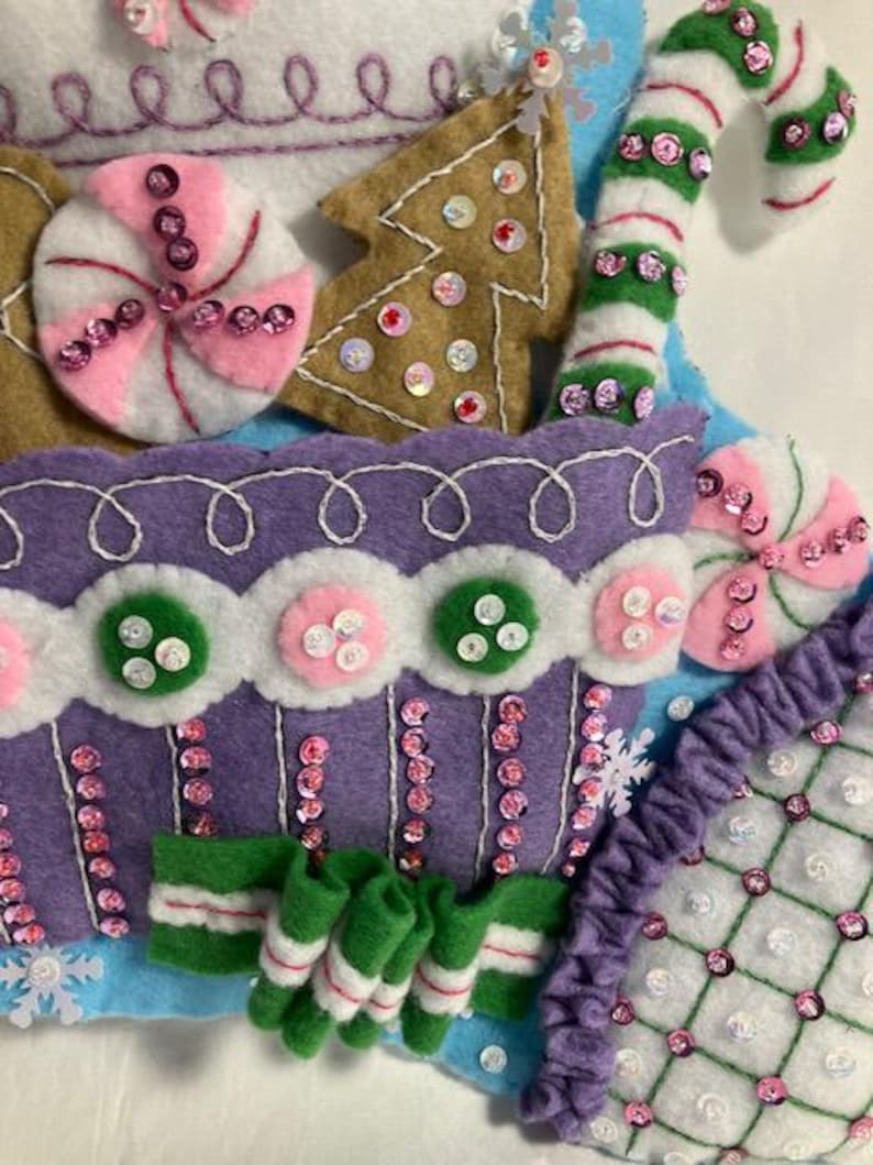 May include: Close-up of a felt Christmas stocking with candy and gingerbread decorations. The stocking features a purple cupcake design, a green and white candy cane, and a gingerbread tree, all embellished with sequins.