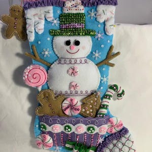 May include: A handmade Christmas stocking with a snowman design. The blue felt stocking features a snowman with a green sequined hat, snowflakes, and various felt candies and gingerbread men. The top is trimmed with pink and purple felt.