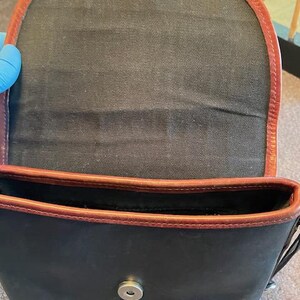 May include: A black canvas bag with brown leather trim and a silver metal snap closure. The bag is open and the inside is visible.
