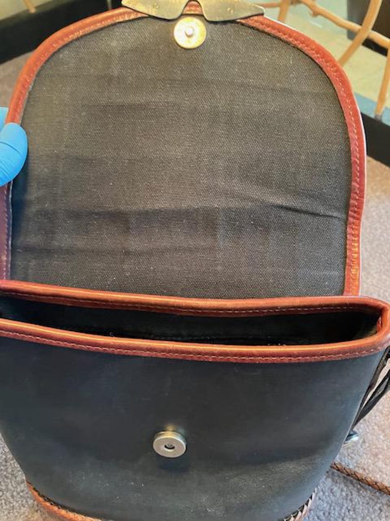 May include: A black canvas bag with brown leather trim and a silver metal snap closure. The bag is open and the inside is visible.