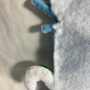 May include: Close-up of a light blue felt object with two blue felt appendages with gold stitching. A white felt appendage with a green accent is also visible. The object is on a white background.