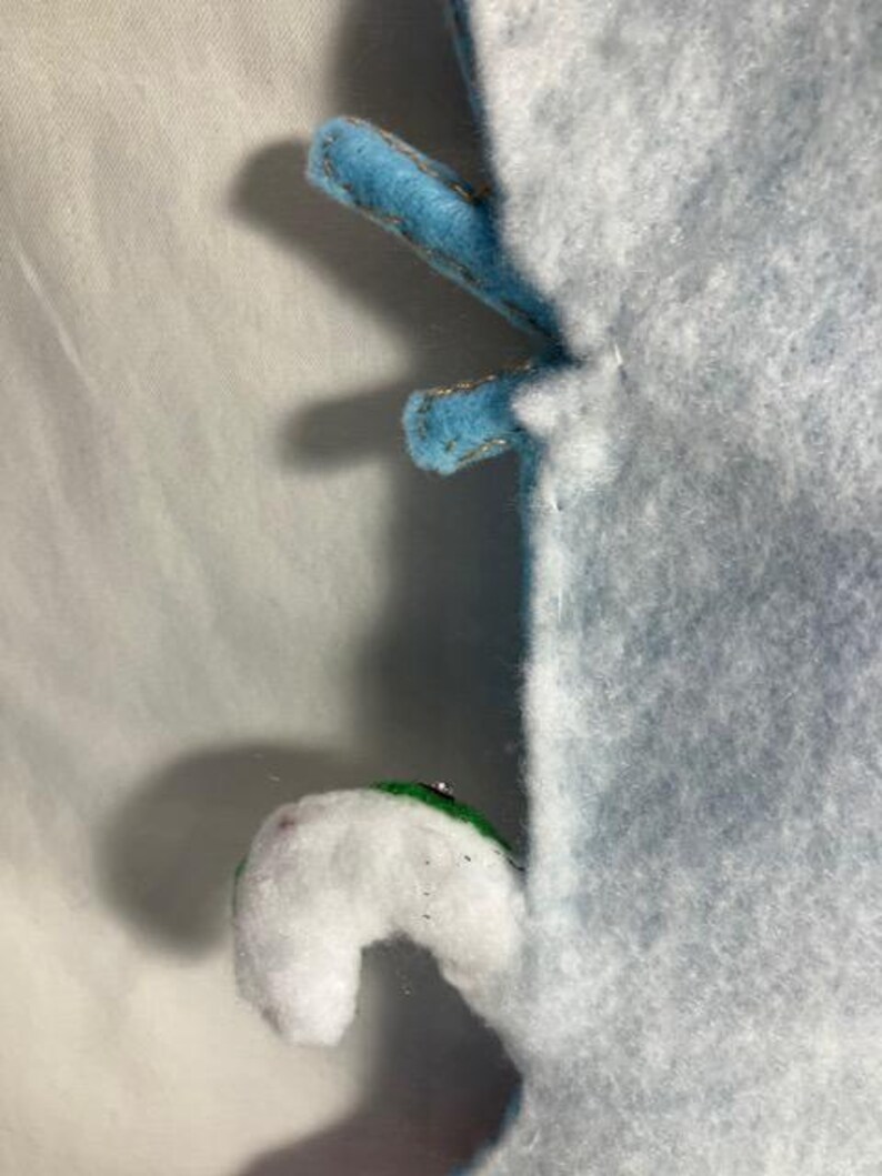 May include: Close-up of a light blue felt object with two blue felt appendages with gold stitching. A white felt appendage with a green accent is also visible. The object is on a white background.