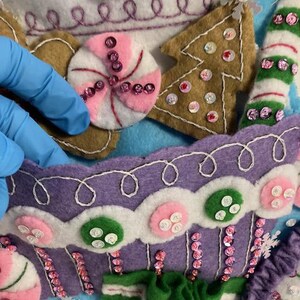May include: Close-up of a felt Christmas stocking with candy and gingerbread house details. The stocking features a purple cupcake design, a gingerbread tree, and a pink and white peppermint candy, all embellished with sequins.