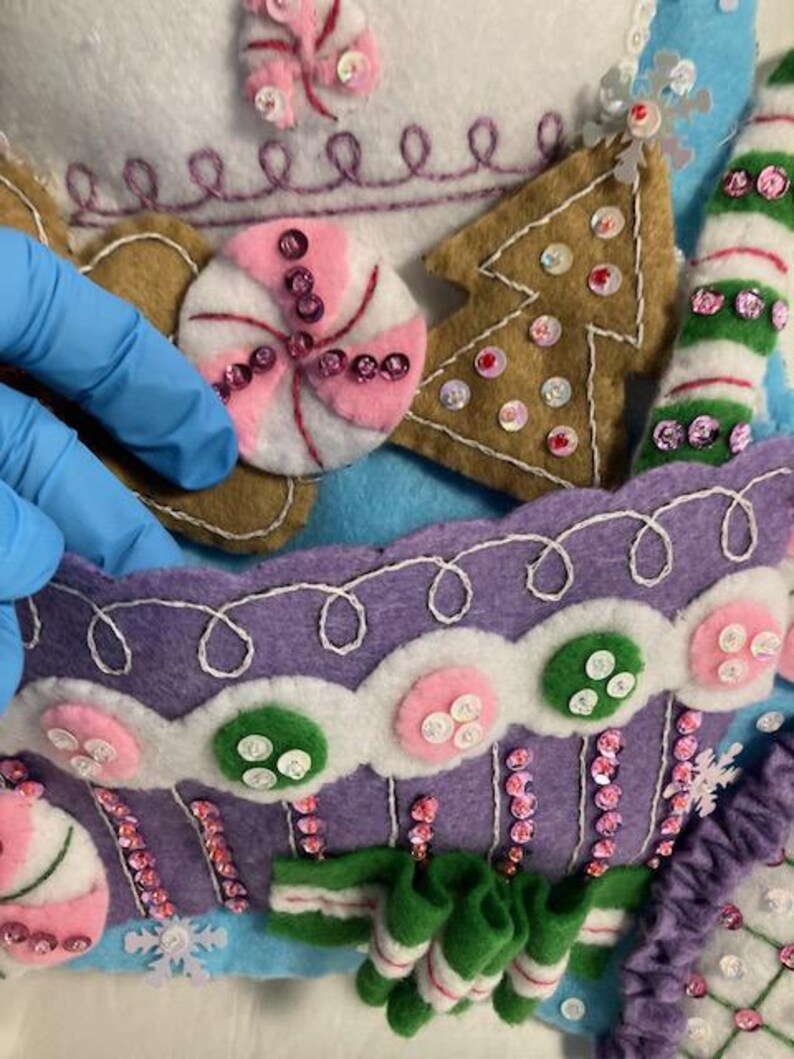 May include: Close-up of a felt Christmas stocking with candy and gingerbread house details. The stocking features a purple cupcake design, a gingerbread tree, and a pink and white peppermint candy, all embellished with sequins.