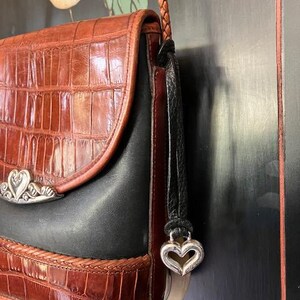 May include: A brown and black leather shoulder bag with a silver heart charm. The bag has a crocodile-embossed pattern and a braided strap.