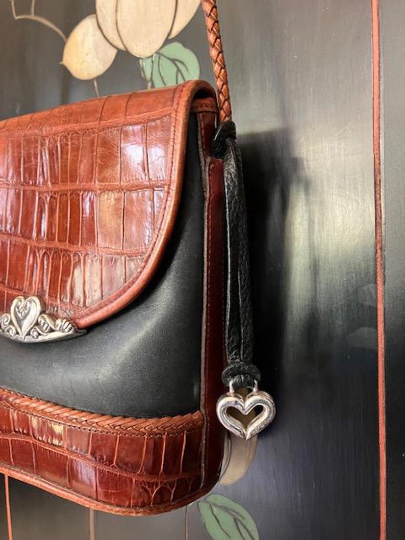 May include: A brown and black leather shoulder bag with a silver heart charm. The bag has a crocodile-embossed pattern and a braided strap.