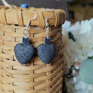 May include: A pair of black lava rock heart earrings with silver arrow charms hanging from silver hooks. The earrings are displayed in a woven basket.