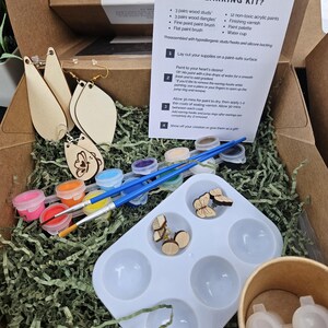 May include: A DIY earring kit with wooden earring shapes, paint, brushes, and a palette. The kit includes instructions and preassembled earring hooks. The box is open, revealing the contents, including a variety of colorful paints.