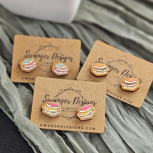 Puede incluir: Tres pares de pendientes con forma de libro sobre tarjetas marrones. Cada juego de pendientes presenta una pila de libros con páginas coloridas. Las tarjetas muestran el texto "Swanger Designs" y "SWANGERDESIGNS.COM".