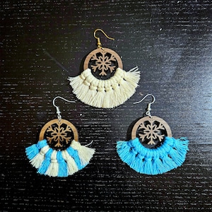 May include: Three pairs of handmade earrings. Each earring features a wooden circular frame with a snowflake cutout. The earrings have a macrame fringe in cream, blue and cream, and solid blue. The earring hooks are gold and silver.