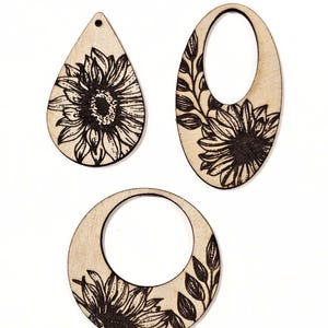 May include: Three wooden earrings with a sunflower design. The earrings are in three different shapes: a teardrop, an oval, and a circle. The sunflower and leaf details are etched in black, creating a contrast against the light wood.