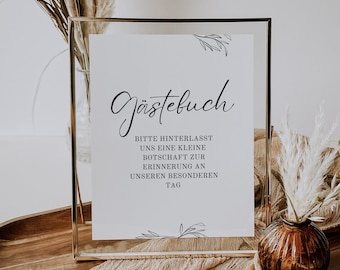 May include: A gold framed sign with the text "Gästebuch" in cursive script. The sign reads "Bitte hinterlasst uns eine kleine Botschaft zur Erinnerung an unseren besonderen Tag". The sign is sitting on a wooden table with a brown vase and dried pampas grass.
