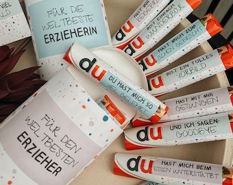 Puede incluir: Una colección de pequeñas barras de chocolate envueltas en papel rojo y blanco con la palabra "du" impresa en ellas. Cada barra tiene un mensaje diferente impreso en ella, como "Du hast mich so viel gelehrt" y "Du hast mich zum lachen gebracht". Las barras están dispuestas en círculo alrededor de un cilindro blanco con una etiqueta azul y blanca que dice "Für die welbeste Erzieherin".
