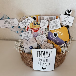 Op de afbeelding: Een gevlochten mand gevuld met diverse items, waaronder een fles, chocolaatjes en reinigingsdoekjes. Verschillende kleine bordjes met Duitse tekst zijn in de mand geplaatst. De bordjes bevatten zinnen als "Anti-Stress" en "Eindelijk pensioen". Witte bloemen zijn gerangschikt.