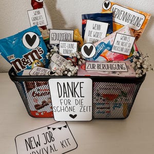 以下が含まれることがあります： M&M's、チョコレート、お菓子などが入った黒いワイヤーバスケット。バスケットには、ドイツ語のテキストとハートのグラフィックが描かれた白いカードがいくつかあります。「New Job Survival Kit」カードが前面にあります。
