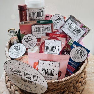 Puede incluir: Una cesta de mimbre llena de artículos de cuidado personal, que incluyen sales de baño, velas, loción corporal, té, parches para los ojos y refrigerios. La cesta está decorada con una etiqueta que dice "Kleine Auszeit für Dich", que se traduce como "Un pequeño tiempo libre para ti".