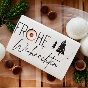 Może przedstawiać: Świąteczna aranżacja bożonarodzeniowa z białym znakiem z napisem "FROHE Weihnachten" i ilustracją drzewa. Scena obejmuje białe bombki, czekoladowe cukierki i gałązki iglaste, wszystko na drewnianej powierzchni.