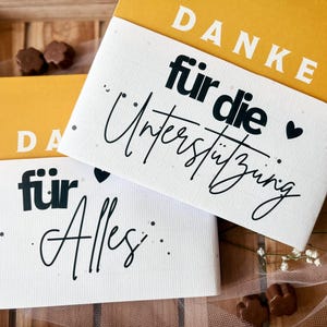 Könnte beinhalten: Zwei Karten mit gelben und weißen Abschnitten. Der obere Teil jeder Karte ist gelb mit dem Wort "DANKE" in Weiß. Der weiße Abschnitt trägt die Wörter "für die Unterstützung" und "für Alles" in schwarzer Schrift. Schokoladenbonbons und Blumen sind um die Karten verstreut.