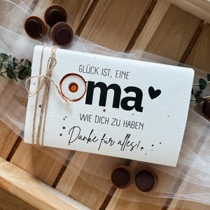 Könnte beinhalten: Ein weißes Holzschild mit deutschem Text, darunter "Glück ist, eine Oma, Wie dich zu haben, Danke für alles!", ist mit Bindfaden gebunden. Das Schild hat ein Herz und ein kreisförmiges Design. Schokoladenbonbons und Grünzeug sind um das Schild angeordnet.