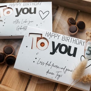 Könnte beinhalten: Zwei weiße Geschenkboxen mit dem Text "HAPPY BIRTHDAY To YOU" und dekorativen Punkten. Eine Box hat ein Herz und den Text "Alles Liebe zum Geburtstag". Die andere Box hat den Text "Lass dich feiern und genie deinen Tag!". Schokoladenbonbons sind verstreut.