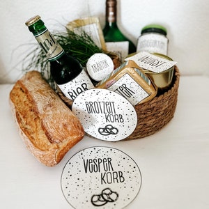 May include: A wicker basket filled with a variety of food and drink items, including a loaf of bread, a bottle of beer, a jar of mustard, a jar of sausage, and a package of butter. The basket is decorated with circular paper tags that say "Brozeit Korb" and "Vesper Korb".