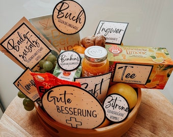 Op de afbeelding: Een houten mand gevuld met beterschapcadeaus, waaronder een boek, thee, gember, citroenen, sap, snoepjes en een kaart met de tekst "Gute Besserung" (Duits voor "Beterschap").