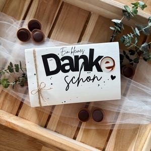 May include: A white wooden sign with the words "Ein kleines Danke sch&ouml;n" in black script and a small heart. The sign is decorated with a twine bow and small black dots. Chocolate candies and greenery surround the sign.