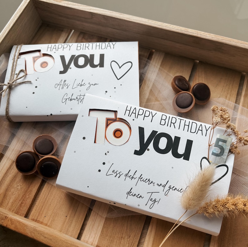 May include: Two white gift boxes with the text "HAPPY BIRTHDAY To YOU" and decorative dots. One box has a heart and the text "Alles Liebe zum Geburtstag". The other box has the text "Lass dich feiern und genie deinen Tag!". Chocolate candies are scattered around.