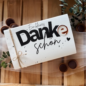 May include: A white rectangular gift box with the words "Ein kleines Danke sch&ouml;n" in black script and a decorative "e" with a brown and white design. The box is tied with twine and decorated with small black dots and a heart.