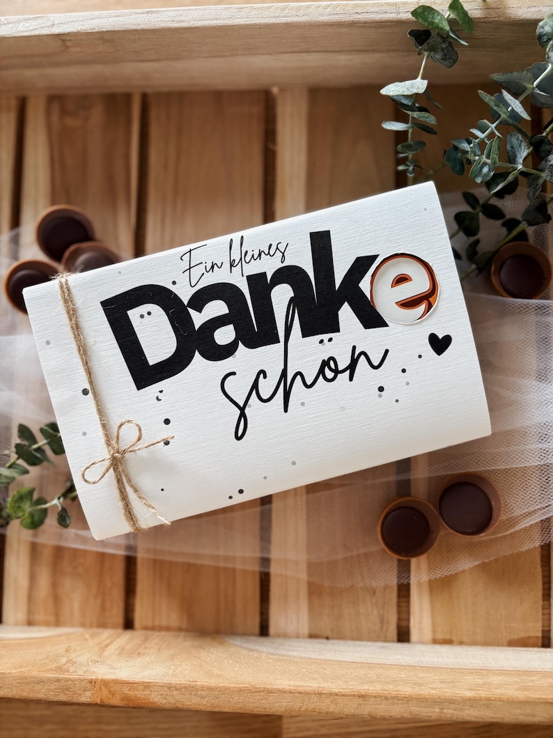 May include: A white rectangular gift box with the words "Ein kleines Danke sch&ouml;n" in black script and a decorative "e" with a brown and white design. The box is tied with twine and decorated with small black dots and a heart.