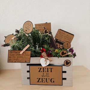Könnte beinhalten: Eine Holzkiste gefüllt mit Grün und bunten Blumen. Die Kiste hat ein Schild mit der Aufschrift "ZEIT statt ZEU". Mehrere kleine Schilder mit Text und herzförmigen Akzenten sind an den Pflanzen befestigt.