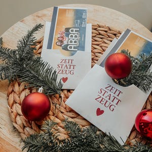 May include: Two white envelopes with the text "Zeit statt Zeug" and a red heart printed on them. The envelopes are decorated with red Christmas ornaments and green pine branches.