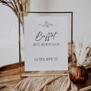 May include: A gold-framed sign with the text "Buffet" in cursive script, followed by "Bitte bedient euch!" and "Guten Appetit". The sign is sitting on a wooden surface with a brown fabric and dried pampas grass.