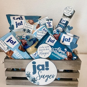 Puede incluir: Una caja de madera llena de varios art&iacute;culos de comida envasados en azul y blanco, incluyendo chocolate, crema de caf&eacute; y galletas. La caja est&aacute; decorada con un c&iacute;rculo grande azul y blanco que dice "ja! Sager".