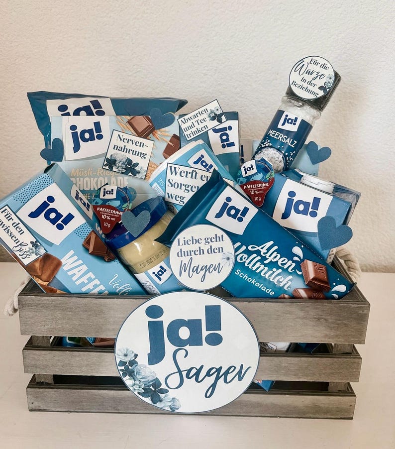 Puede incluir: Una caja de madera llena de varios art&iacute;culos de comida envasados en azul y blanco, incluyendo chocolate, crema de caf&eacute; y galletas. La caja est&aacute; decorada con un c&iacute;rculo grande azul y blanco que dice "ja! Sager".