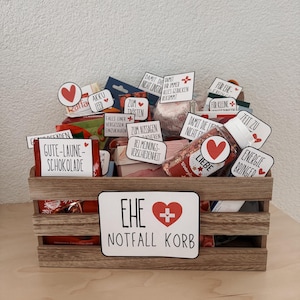 Pode incluir: Uma caixa de madeira cheia de vários itens, incluindo chocolates, doces e pequenos presentes. A caixa tem vários rótulos com texto em alemão e um sinal que diz "EHE NOTFALL KORB" com um coração e uma cruz. A caixa está em uma superfície de madeira clara.