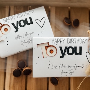 May include: Two white birthday gift boxes with the words "HAPPY BIRTHDAY TO YOU" in black and orange. One box is tied with twine. The boxes are on a wooden surface with chocolate candies.