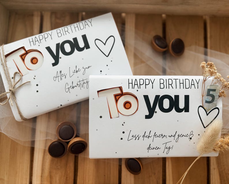 May include: Two white birthday gift boxes with the words "HAPPY BIRTHDAY TO YOU" in black and orange. One box is tied with twine. The boxes are on a wooden surface with chocolate candies.