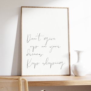 Könnte beinhalten: Ein gerahmter Kunstdruck mit dem Text "Don't give up on your dreams. Keep sleeping." in eleganter Schreibschrift. Der Druck hat einen hellen Hintergrund und einen Holzrahmen. Eine weiße Vase und ein Stuhl sind ebenfalls zu sehen.