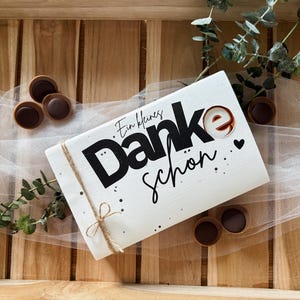 May include: A white rectangular gift box with the words "Ein kleines Danke sch&ouml;n" in black script and a brown "e" is tied with twine. The box is surrounded by chocolate candies and greenery, all on a wooden tray.