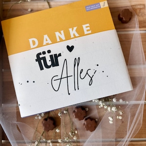 May include: A gift box with the words "DANKE f&uuml;r Alles" in white and black lettering on a yellow and white background. The box is surrounded by chocolate flower-shaped candies and white flowers.