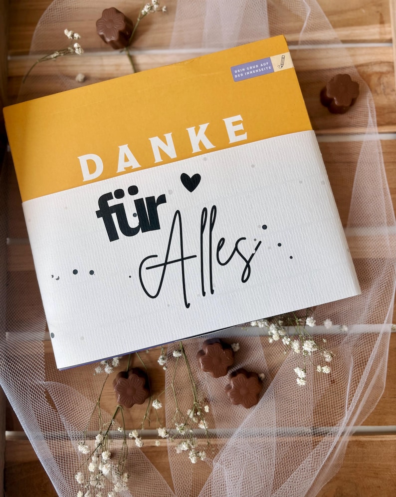 May include: A gift box with the words "DANKE f&uuml;r Alles" in white and black lettering on a yellow and white background. The box is surrounded by chocolate flower-shaped candies and white flowers.
