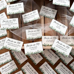 May include: A collection of white cards with black script and green floral accents, arranged on a wooden surface. The cards feature phrases in German, suggesting a wedding or event theme. Some cards are visible with phrases like "Mache ein Foto".