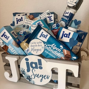 Puede incluir: Una caja de madera llena de varios art&iacute;culos de comida, incluyendo barras de chocolate, una caja de gofres, una botella de sal y un tarro de crema de caf&eacute;. La caja est&aacute; decorada con una gran letra blanca "J" y un c&iacute;rculo azul y blanco con el texto "ja! Sager".
