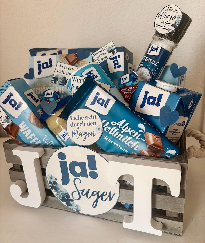 Puede incluir: Una caja de madera llena de varios art&iacute;culos de comida, incluyendo barras de chocolate, una caja de gofres, una botella de sal y un tarro de crema de caf&eacute;. La caja est&aacute; decorada con una gran letra blanca "J" y un c&iacute;rculo azul y blanco con el texto "ja! Sager".